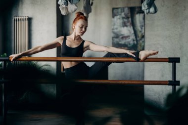 Esneklik üzerine çalışıyorum. Genç kızıl saçlı kadın balerin bir bacağını kaldırdı ve stüdyonun ya da sınıfın karanlık geçmişine karşı esneme egzersizleri yaptı. Barre egzersiz konsepti