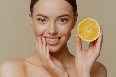 Güzel, narin bir kadının yakın çekimde gülümsemesi sağlıklı, bakımlı bir cilde sahip limon dilimleri güzellik için narenciye ve kahverengi arka plan üzerinde üstsüz duran sağlık tezgahları kullanır.