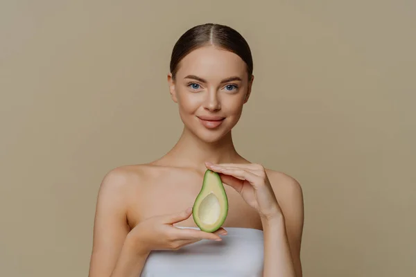 Hygiene skin care and facial treatments concept. Indoor shot of pleased female model holds half of avocado wrapped in bath towel going to use beauty product nourishes skin isolated over beige wall