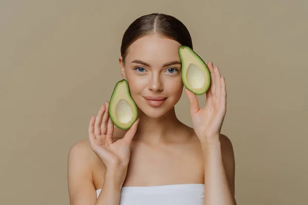 Portrait of young woman with dark combed hair relaxes in bath with avocado wears minimal makeup wrapped in towel has minimal natural makeup isolated over brown background. Skin care concept.