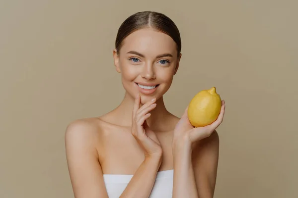 Cosmetology skin care and beauty concept. Pleased brunette young woman wrapped in bath towel holds lemon for making facial mask has refreshed clean skin after taking shower and spa procedures