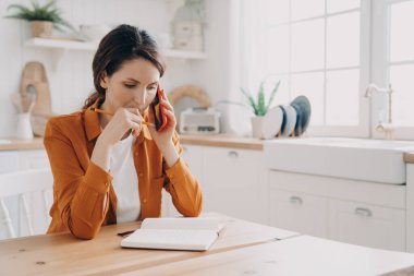 Şaşkın kadın telefon ediyor, mutfak masasında oturmuş notlar alıyor. Düşünceli kadın serbest çalışan müşteriye danışmanlık yapıyor ya da evdeki meslektaşımla projeyi tartışıyor. Uzak görev konsepti.