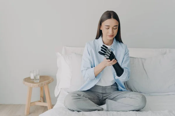 Modern girl with disability training to install her bionic prosthetic arm, sitting on bed at home. Young woman using high tech prosthesis after limb loss. Advertising of robotic artificial limbs.
