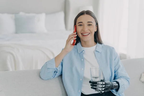 Smiling young caucasian disabled girl answer call, speak by phone using artificial limb, bionic arm prosthesis, holding smartphone and glass of water, sitting on sofa at home. Medical high tech.