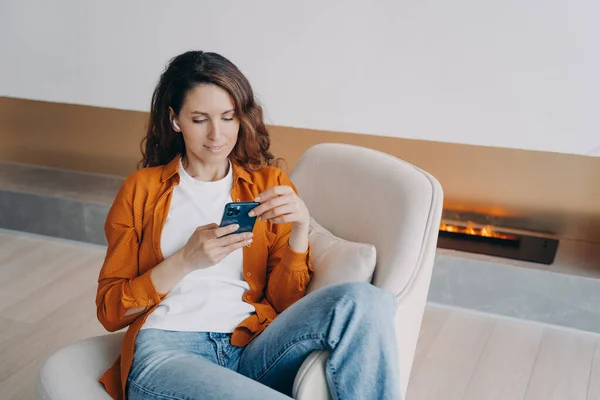 Modern hispanic woman in wireless earphones using smartphone apps, choosing music playlist, audio book, sitting in armchair. Female browsing social networks, shopping online, listening to new podcast.