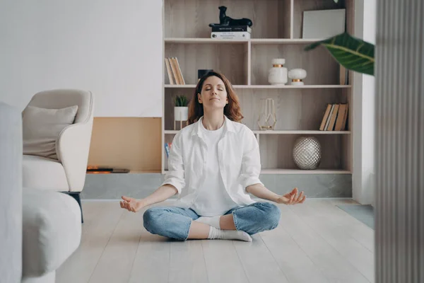 Calm woman sitting in lotus position with mudra gesture on floor in modern living room. Peaceful female meditates, practicing yoga, breathing fresh air alone at home. Recreation, healthy lifestyle.