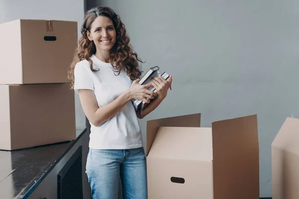 Smiling spanish female preparing moving to new home packing things in cardboard boxes. Glad woman holding stack of notebooks looking at camera on relocation day. Real estate sale, dwelling rental.