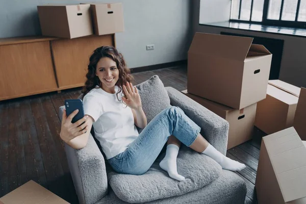 Happy female homeowner blogger holding smartphone greeting followers waving hand, sitting near cardboard boxes with belongings on relocation day to new home. Moving and repair concept.