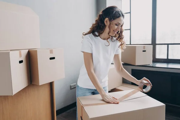 Spanish woman packing things in cardboard box using adhesive tape in empty room or storage. Young female preparing to move or packs parcel in carton. Delivery service or moving company concept.