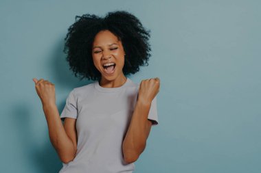 Neşeli kız yumruklarını mutlulukla sıkar, yüksek sesle bağırırken ağzını açar. Afro-Amerikalı kızın fotoğrafı mavi stüdyo geçmişinde günlük kıyafetler giyiyor. Duygular ve olumlu duygular kavramı