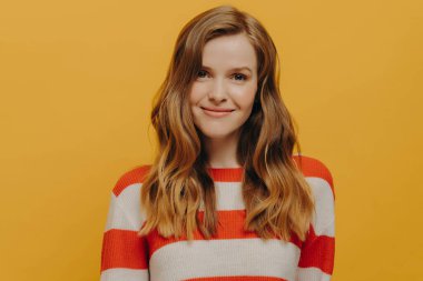 Uzun dalgalı kahverengi saçlı, sıcak bir gülümsemeyle kameraya bakan, harika hisseden ve sarı arka planda izole bir şekilde duran, günlük süveter giyen nazik bir kadın.