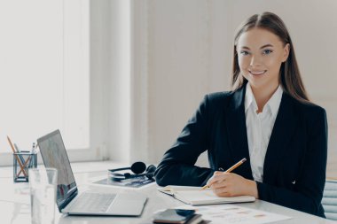 Siyah takım elbiseli, gülümseyen beyaz bir kadının portresi. Kameraya bakıyor. Not defteri ve dizüstü bilgisayarının yanında oturuyor..