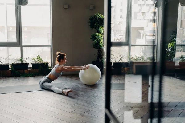 Side view of young red haired woman with perfect slim body sitting in split position on yoga mat in fitness studio bending forward and touching large stability exercise ball next to panoramic window