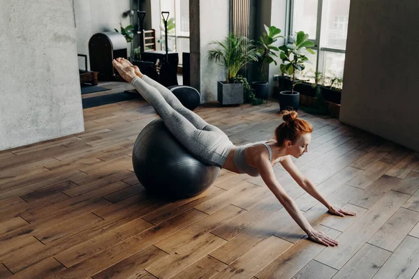 Ball workout for balance and stability. Young sportive woman lying on fitball with legs squeezed together and directed upwards while supporting body with straight arms based on floor. Fitness concept