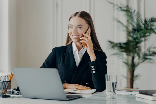 Young cheerful professional businesswoman in formal suit talking on phone, smiling and discussing business ideas with copartner, happy female office worker sitting in front of laptop behind work desk