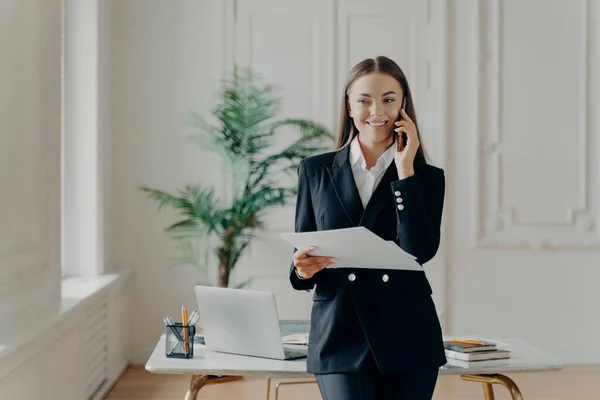 Professional young business lady in formal suit looking away while talking on phone and holding notes, business leader laughing when hearing good news, happy female office worker standing at office