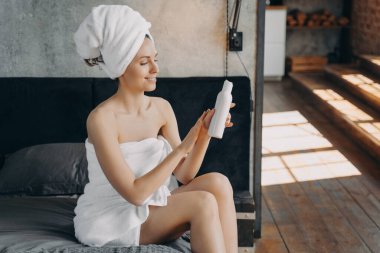 Havluya sarılmış bir kadın elinde güneş kremi şişesiyle otelde yatakta yatarken duştan sonra kozmetik kullanıyor. Gülümseyen kadın nemlendirici cilt, tatilde cilt bakımı rutini. Vücut bakımı