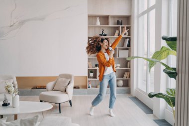 Enerjik bir kadın akıllı telefon tutuyor, eğleniyor, modern oturma odasında dans ediyor, telefonda müzik eşliğinde heyecanla dans ediyor, şarkı söylüyor, evde boş zaman geçirmekten keyif alıyor..