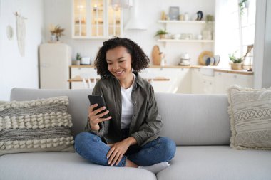 Gülümseyen Afrikalı Amerikalı genç kız sosyal ağda sohbet ediyor, modern cep telefonu uygulamaları kullanıyor, evdeki rahat koltuğunda oturuyor. Akıllı telefon tutan, online alışveriş yapan mutlu melez genç bayan..