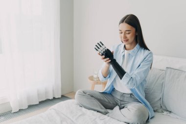 Gülümseyen beyaz engelli kız yapay koluna dokunuyor, evde yatakta oturuyor. Biyonik protez eli özelleştiren memnun bir kadın. Engelli insanlara yüksek teknoloji bakımı.