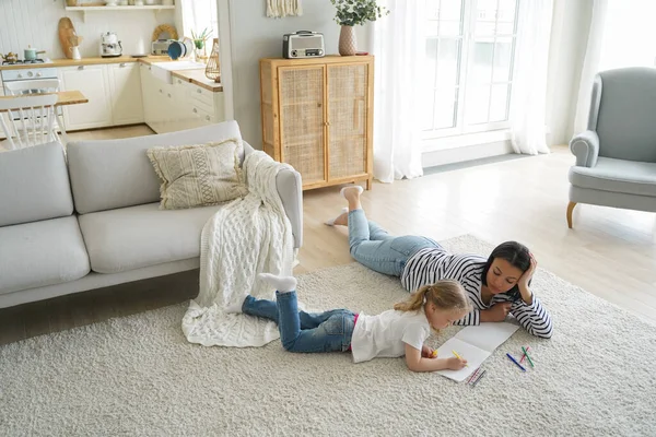 European mom is teaching kid how to draw. Family weekend at home. Young mother and little girl lying on floor together and painting with colorful markers. Woman and child have leisure in apartment.