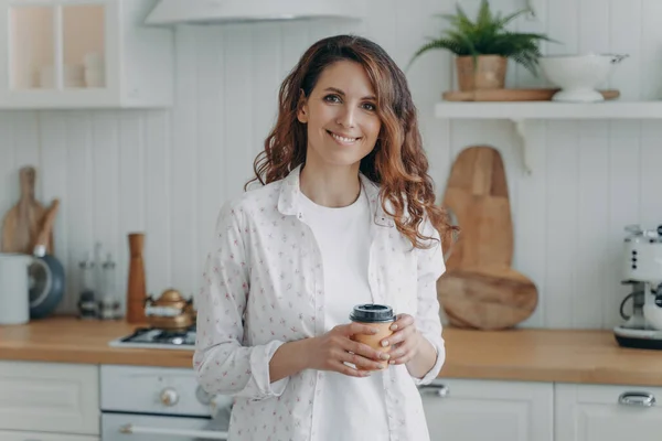 Relaxed smiling hispanic woman with paper cup of coffee standing in cozy modern kitchen, looking at camera. Pleased happy female homeowner or tenant housewife enjoying domestic routine at home.