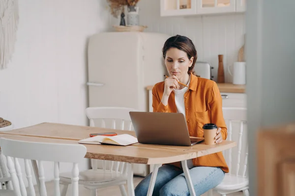 家庭からオンラインで働く ノートパソコンの画面を見つめてキッチンに座っている普及した女性は 解決策やアイデアを検索します プロジェクトを検討している女性のフリーランス労働者のビジネス女性の思考 リモートジョブ — ストック写真