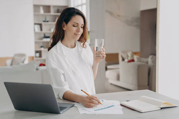 Businesswoman working at laptop, notes, drinking glass of pure water at workday beginning. Female manager enjoy healthcare morning daily routine at home office. Remote job, healthy lifestyle