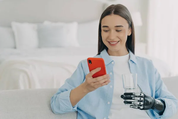 Smiling young disabled woman holding smartphone and glass of pure water. Female with bionic prosthetic arm using mobile phone apps, chatting in social networks, shopping online at home.