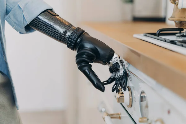 Bionic prosthetic arm adjusting temperature on gas hob control panel, person with disability using stove by artificial robotic hand, close-up. Domestic life routine and people with disabilities.