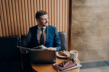 Başarılı bir şekilde gülümseyen mavi takım elbiseli bir iş adamı, elinde gazete tutarken dizüstü bilgisayarla kafede oturup kruvasanla kahve ısmarlıyor.