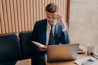 Zarif bir finansör, kulaklıkla düşünceli bir görünüm ve iş sunumunda internette çalışırken bir fincan latte içerek ve çalışma kitabındaki notlarını kontrol ederek.