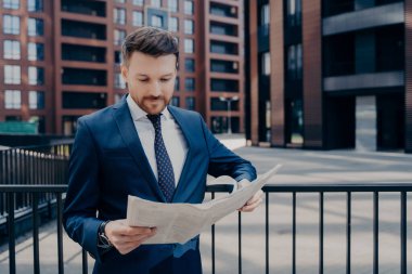 Odaklanmış iş adamı takım elbise giymiş ilginç haberler için son gazeteleri kontrol ediyor, dışarıda tek başına dikilip ofis binalarının yanındaki siyah çitlere yaslanıyor, dışarıda çalışmaktan keyif alıyor.