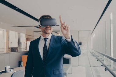 Üç boyutlu görselleştirme için vr gözlük kullanan gülümseyen erkek girişimci, parmağıyla işaret eden, sanal gerçeklik gözlüğü takan iş adamı, modern ofiste dururken dijital dünyayla etkileşim halinde olan