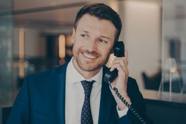 Yeni aldığı terfiyi bildirmek için karısını arayan tatmin olmuş erkek ofis çalışanı. Kirli sakallı, parlak gülüşlü, ofisin arka planında bulanık duran genç yakışıklı bir adam.