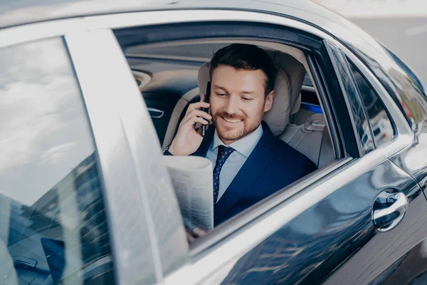 Positive young company executive reading latest newspaper, sharing good news regarding with colleague or partner, satisfied with publicity while riding in back of car with open window