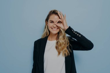 Uzun sarı saçlı, siyah ceketli ve beyaz gömlekli mutlu bir genç kadın resmi. Elleriyle iyi bir jest yapıyor ve parmaklarından bakıyor. Güzel bir gün geçiriyor ve ne kadar harika olduğunu gösteriyor.