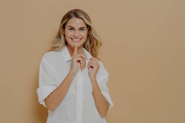 Gizlilik kavramı. Genç, neşeli, güzel bir kadın, işaret parmağını dudaklarında tutuyor ve sessiz bir jest yapıyor. Bej arka planda fotokopi aletiyle poz verirken sessiz olmak istiyor.