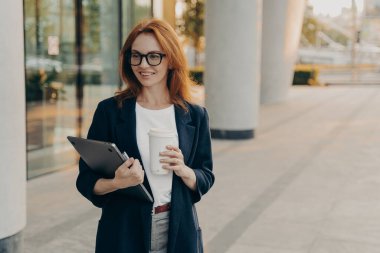 Ofisine giderken dizüstü bilgisayar ve bir fincan kahve taşıyan mutlu Avrupalı kızıl saçlı iş kadını şehir merkezinde açık havada yürüyen bir bayan girişimci. Stil sahibi serbest insanlar.