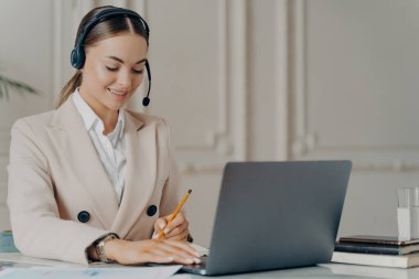 Dost canlısı, kulaklık takan, mikrofonlu bir kadın CEO 'nun portresi dizüstü bilgisayardaki web konferansına katılıyor, resmi takım elbiseli genç bir iş kadını iş yerinde otururken notlar yazıyor.