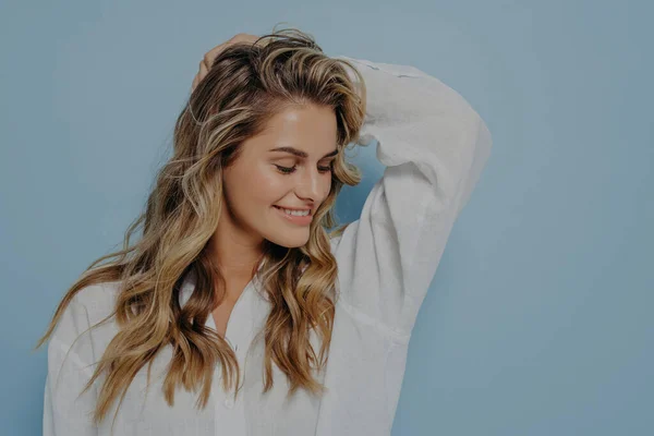 Relaxed romantic blonde woman with long wavy hair and hand behind her head, looking down with bright smile, dressed in white shirt with unbuttoned top while standing alone next to light blue wall