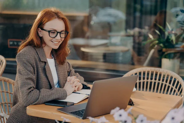 Redhead female freelancer watches training webinar makes notes in notepad sits in front of opened laptop computer dressed formally poses at outdoor cafe browses internet develops new project