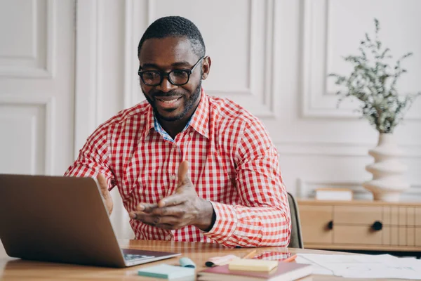 积极的非洲男子在线教师导师看着笔记本电脑屏幕 面带微笑 一边与学生交谈 一边从家里远程工作 穿着休闲装 电子学习和远距离教育 — 图库照片
