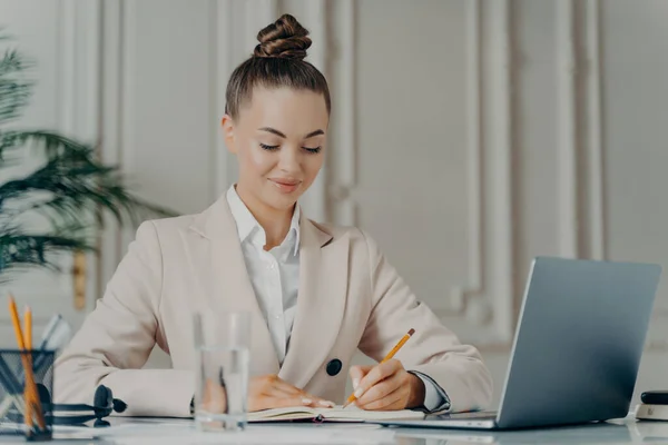 Making to do list. Successful businesswoman in elegant suit writing down points in notepad while sitting at home office, planing working day. Happy female employee in formal wear enjoying work