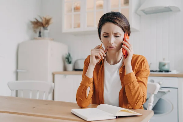 Nervous frowning woman making phone call customer service, having unpleasant conversation, sitting at table with notebook in kitchen. Businesswoman freelancer working at home, discussing project.