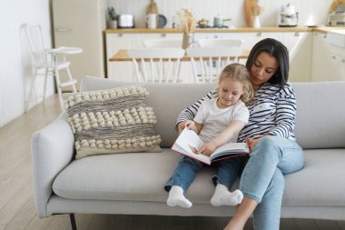 Genç anne ve evlat edinilmiş anaokulu kızı birlikte kitap okuyor, oturuyor ve evdeki kanepede sarılıyorlar. Anne küçük kızı eğitiyor, kanepede masal okuyor. Annelik, evlat edinme kavramı.