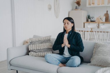 Sakin, genç bir kadın evdeki kanepede oturup yoga yapıyor. Kadın, kanepede nilüfer çiçeği duruşunda namaste jesti yaparak meditasyon yapıyor. Sağlık, stres rahatlama konsepti.