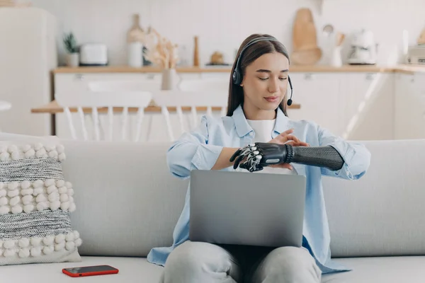 Young disabled girl freelance employee in headset working at laptop online, sitting on sofa at home, setting her bionic prosthetic arm. People with disabilities and daily routine concept.