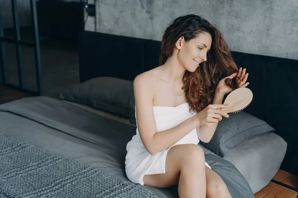 Gorgeous european woman combs her long silky hair in bedroom. Hair cosmetic and hair treatment after bathing. Young woman takes morning shower at home and doing hair. Femininity and beauty concept.