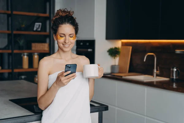 Young woman applies anti wrinkle eye patches and relaxing at home texting on phone. Happy hispanic girl wrapped in towel after spa procedures. Body care, beauty and wellness. Modern interior.